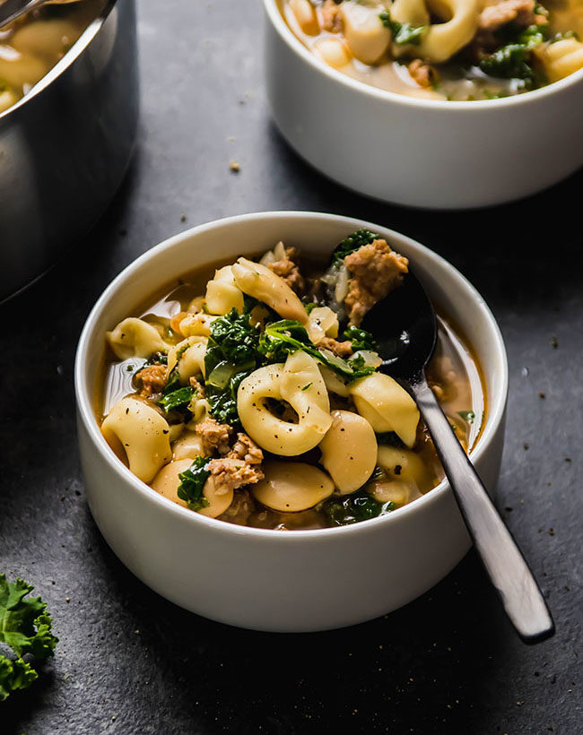 Tortellini soup with sausage and kale in a white bowl with a black spoon.