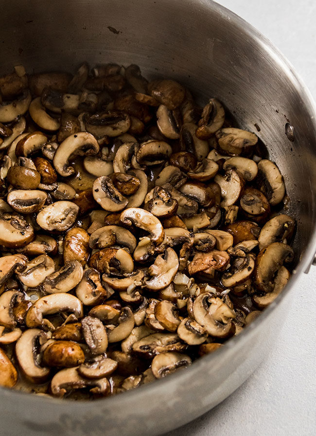 Sautéed mushrooms and garlic in a silver saucepan on a white counter.