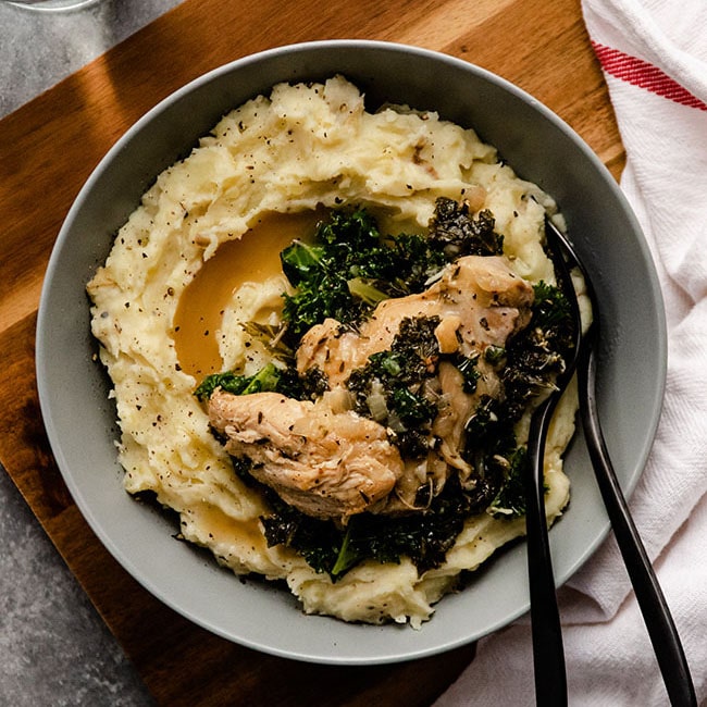 Chicken thighs, kale, and gravy over mashed potatoes in a shallow blue bowl.