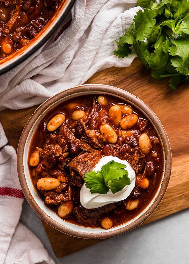 Beef chili with white beans in a brown bowl, topped with cilantro leaves and sour cream.