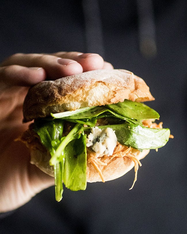 Man\'s hand holding a buffalo chicken sandwich with arugula in front of a dark background.