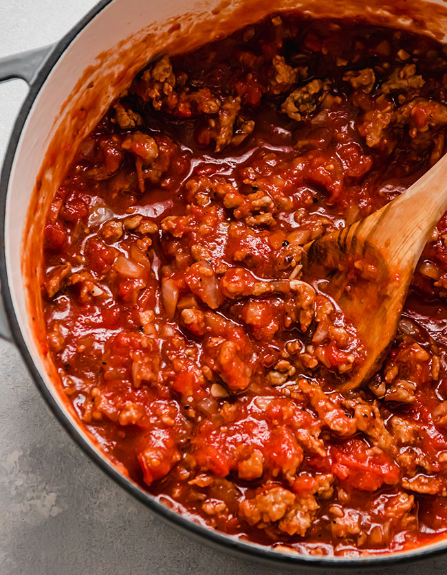 Wooden spoon stirring Italian sausage and tomato sauce together in a grey pot.