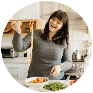 Jessie standing in a white kitchen holding a fork with pasta twirled around it.