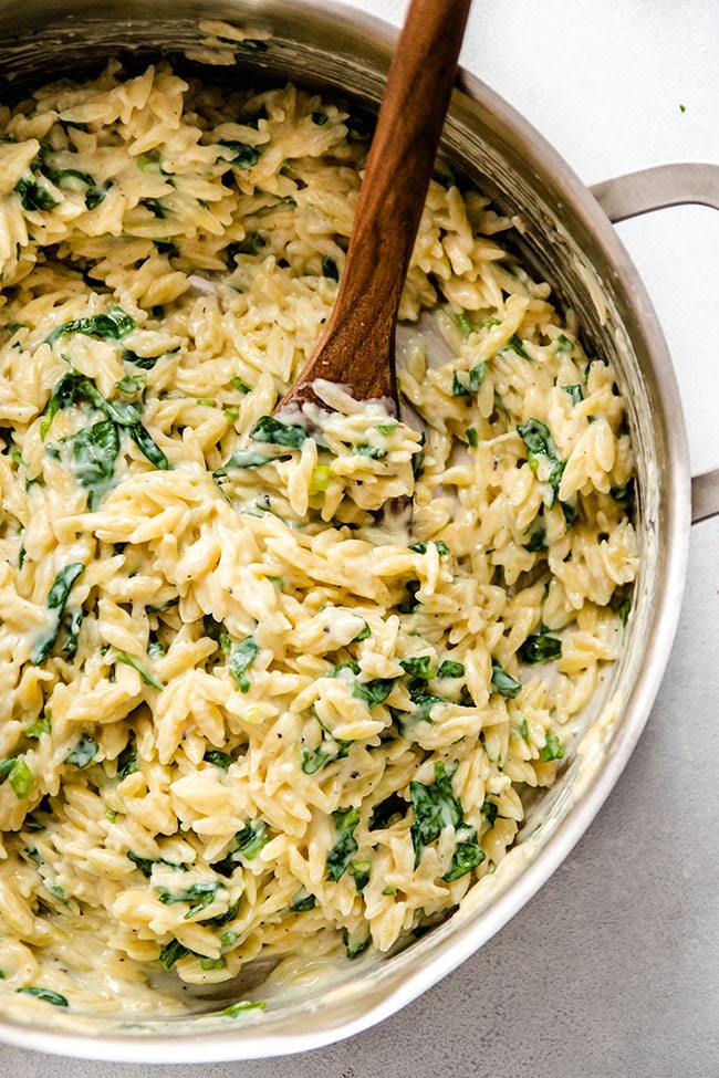 Wooden spoon stirring orzo and spinach cream sauce together in a silver pot.