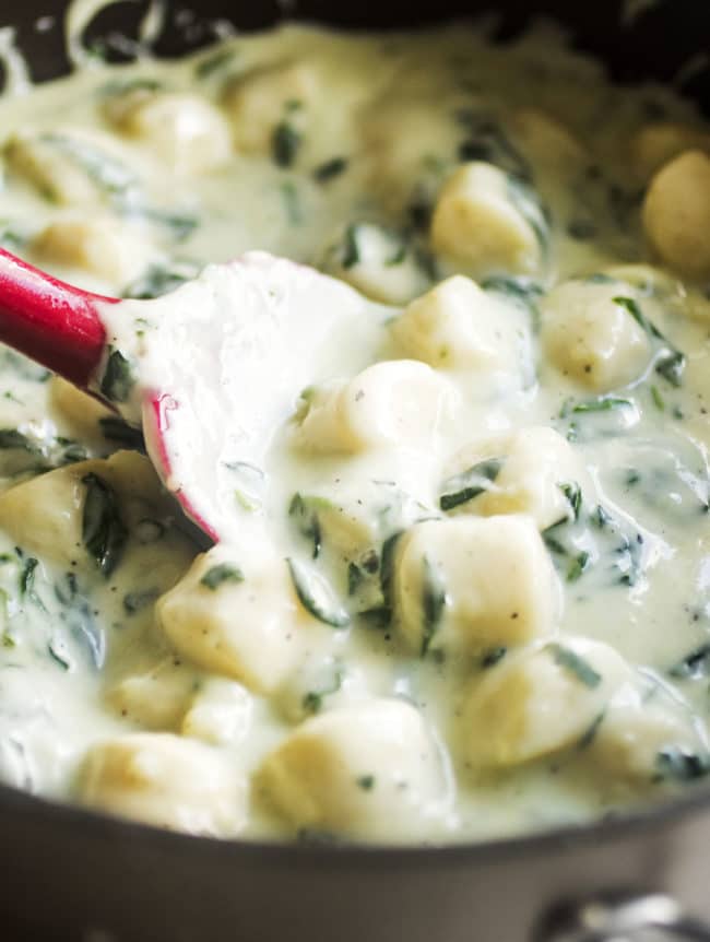 Red spatula stirring gnocchi with cream sauce in a large pot.