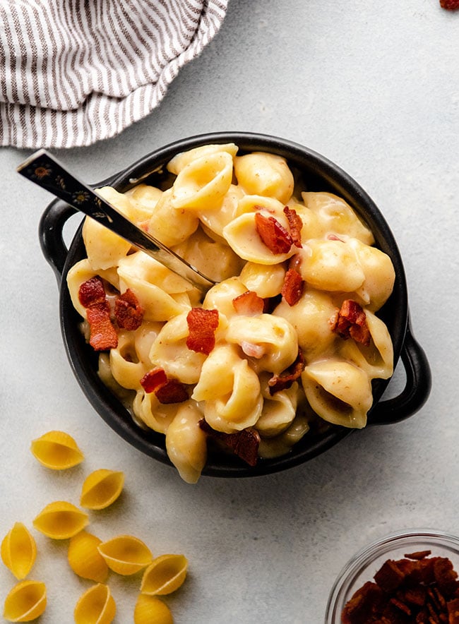 Spoon stirring shell pasta and crispy bacon in a black baking dish.