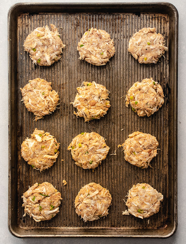 Twelve uncooked crab cakes arranged on a large baking sheet.