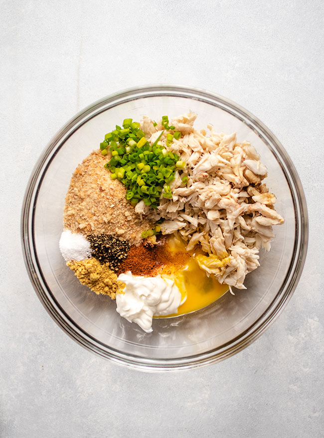 Glass bowl filled with crab, bread crumbs, egg, mayonnaise, spices, and green onions.