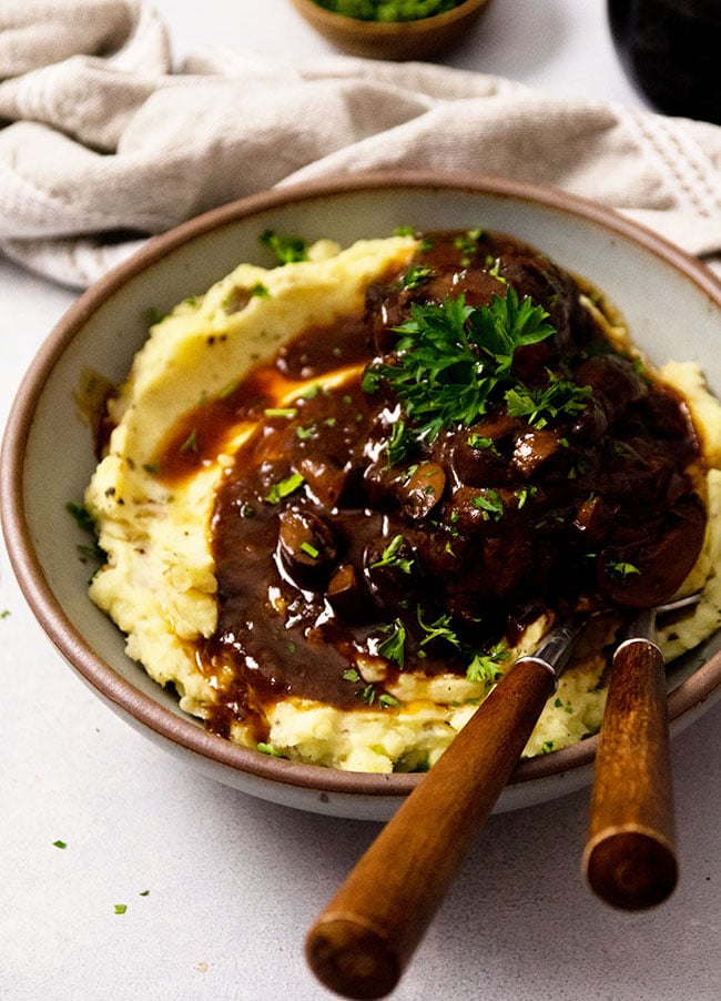 Two wooden forks on the side of a grey bowl filled with mashed potatoes and short ribs.