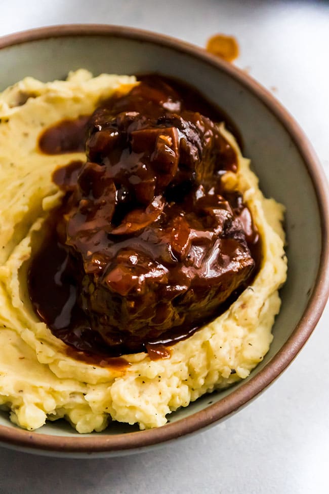 Mashed potatoes topped with short ribs and a red mushroom sauce in a grey bowl.