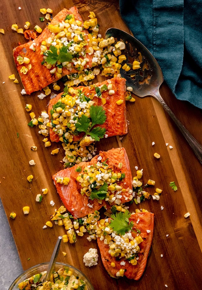 A sockeye salmon fillet on a wooden cutting board, topped with grilled corn salsa and fresh cilantro.