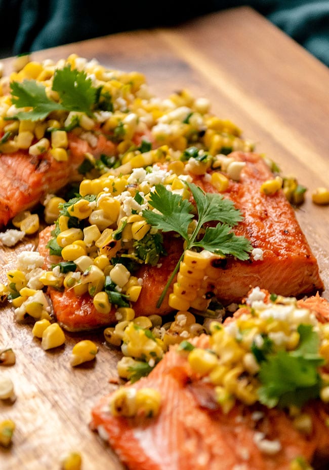Grilled sockeye salmon portion topped with grilled corn salsa and fresh cilantro on a wood surface.