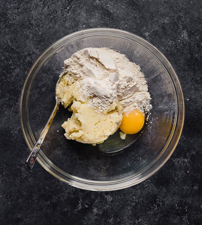Mashed potatoes, flour, and an egg in a glass bowl on a black countertop.