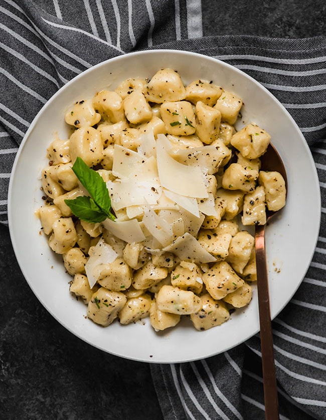 Potato gnocchi in a white bowl atop a striped napkin, topped with parmesan cheese and fresh basil.