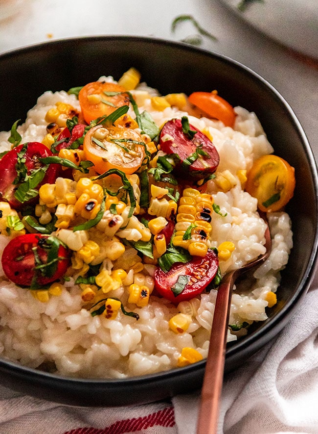 Risotto topped with halved cherry tomatoes, grilled corn, and fresh basil in a black bowl with a copper fork.