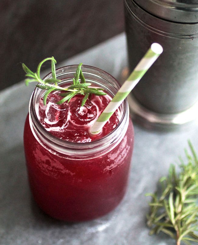 Bright pink blackberry cocktail in a glass mason jar with a sprig of fresh rosemary and a green and white striped straw.