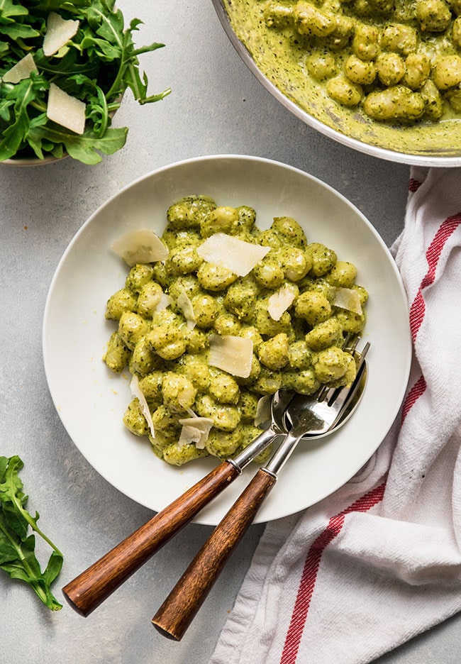 A white bowl filled with gnocchi, tossed with pesto cream sauce and shaved parmesan cheese.