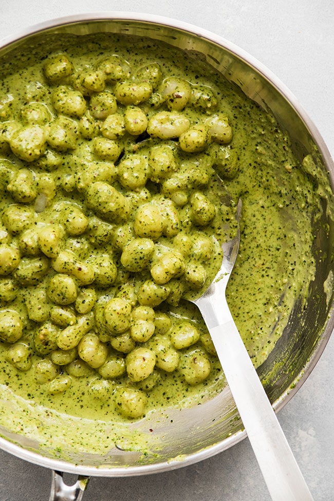 Metal spoon stirring fresh gnocchi with pesto and heavy cream in a large skillet.