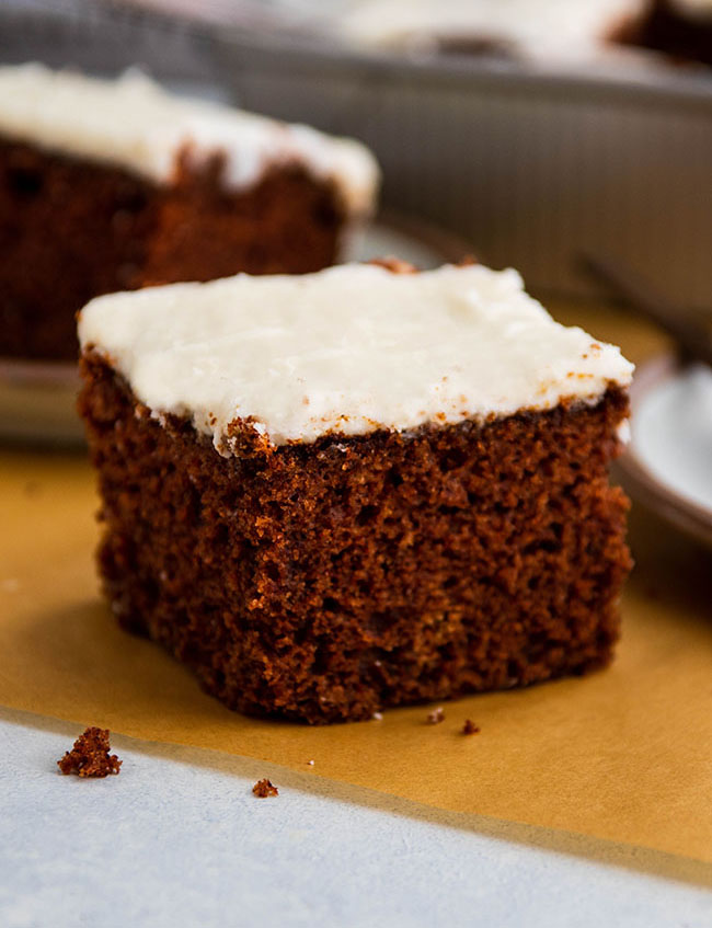 Square piece of brown cake with white frosting, sitting on a piece of brown parchment paper.