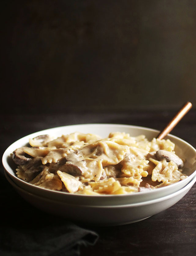 Shallow white bowl filled with bowtie pasta and a white mushroom sauce on a dark brown table.