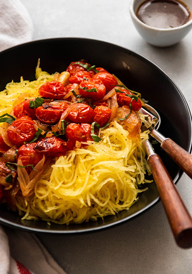 Two wooden forks in a shallow black bowl filled with shredded spaghetti squash and roasted tomatoes.