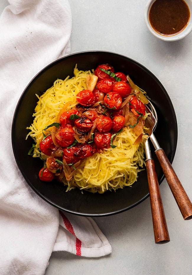 Spaghetti squash strands topped with roasted tomatoes in a shallow black bowl with two wooden serving forks.