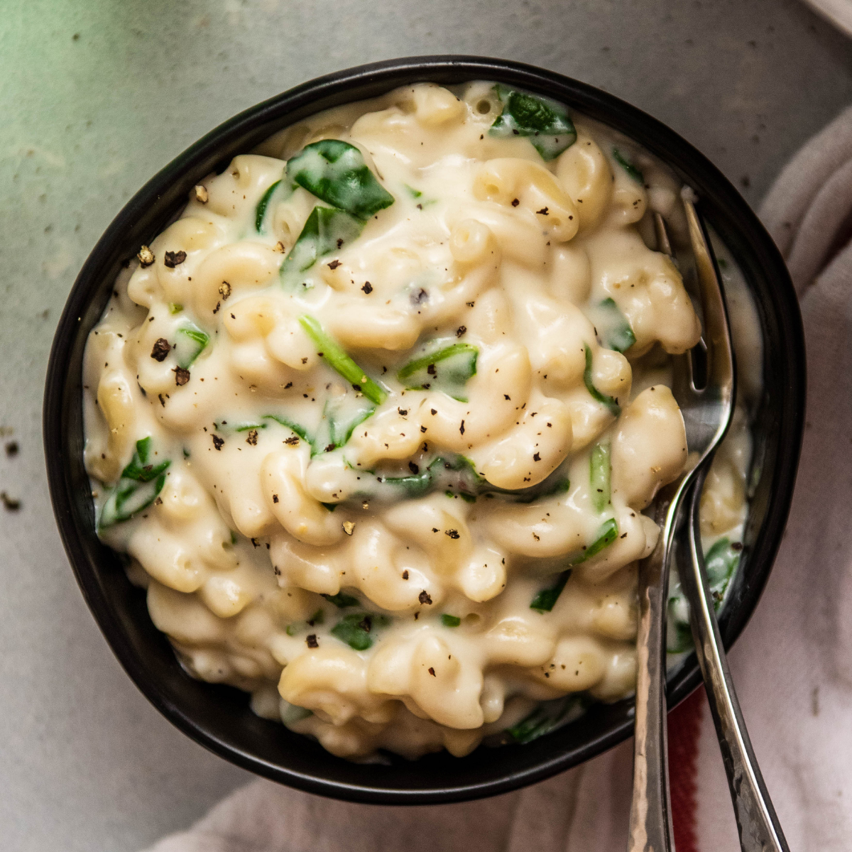Macaroni with white sauce and spinach in a black ceramic bowl.