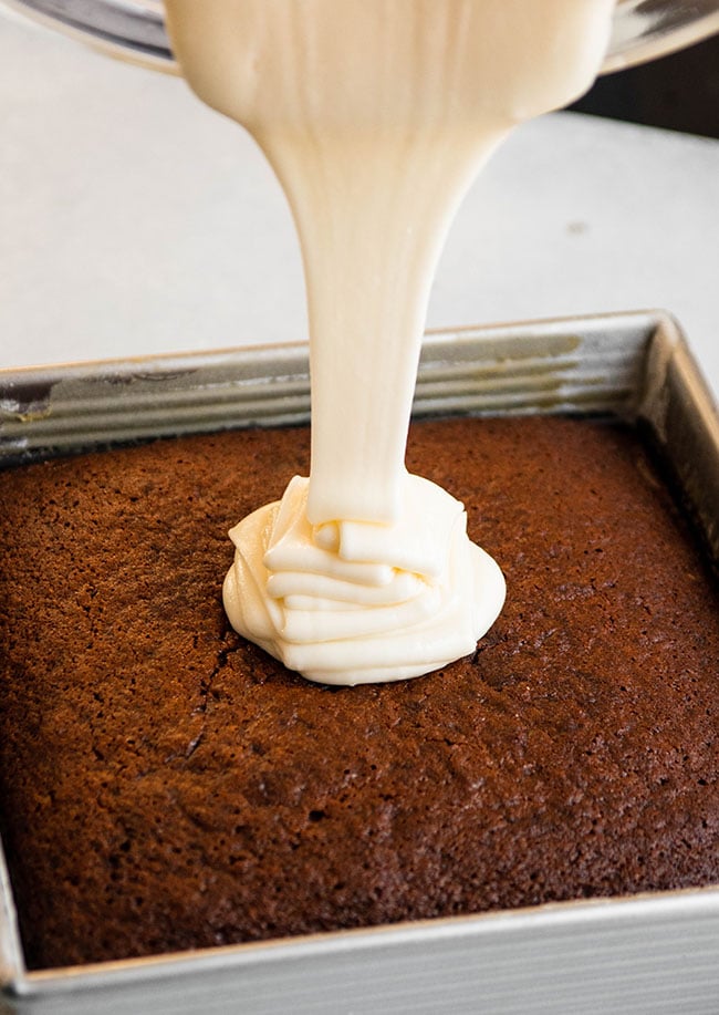White frosting being poured onto a brown cake in a square pan.