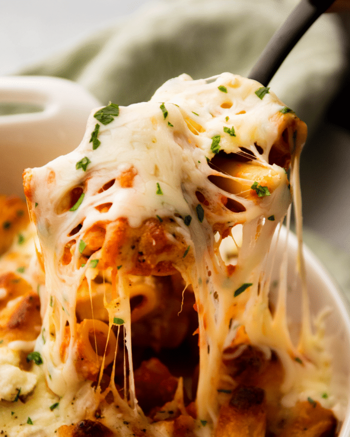 Spoon lifting baked ziti out of a white baking dish.