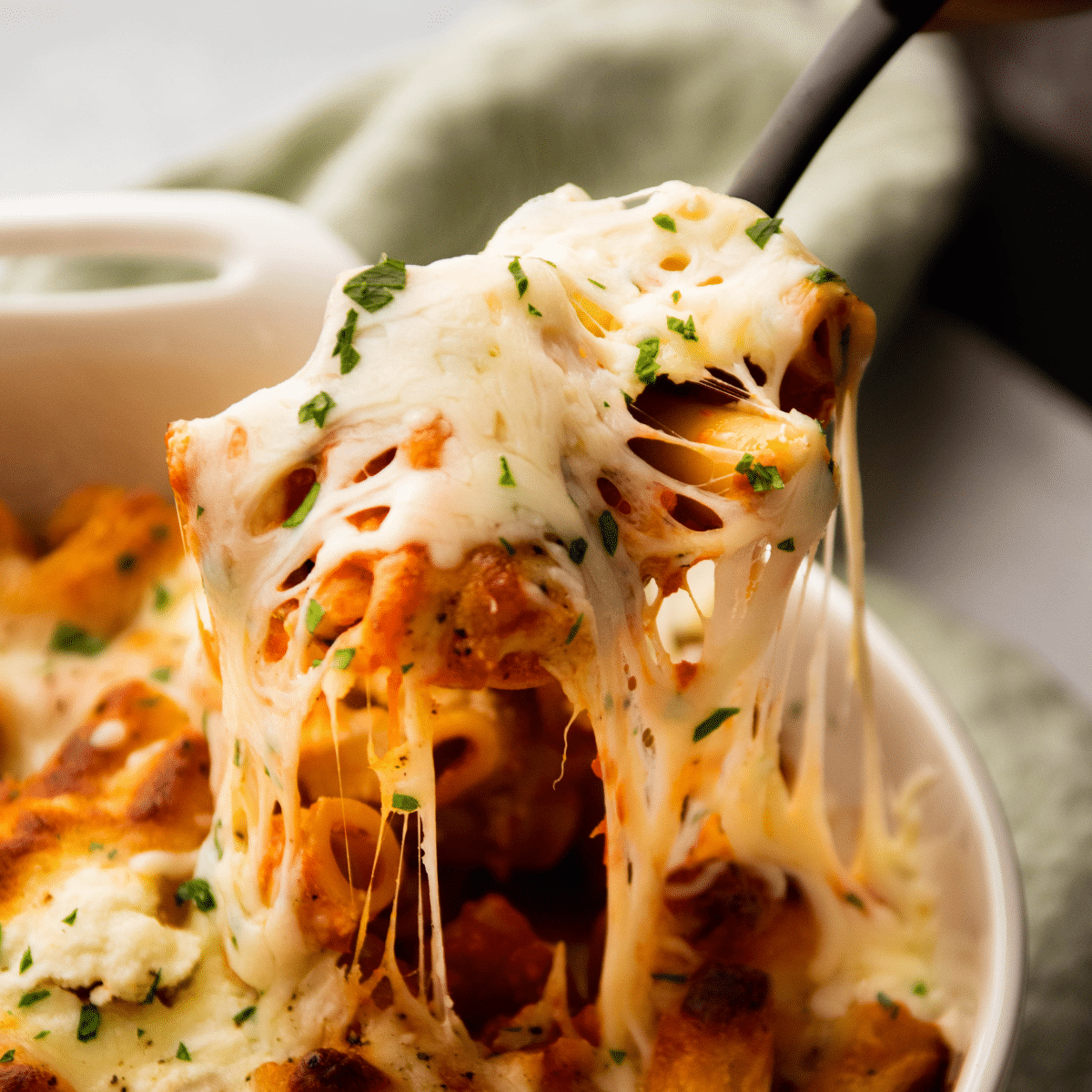 Spoon lifting baked ziti out of a white baking dish.