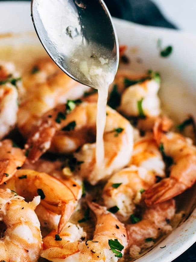 Silver spoon pouring white wine garlic sauce over baked shrimp.