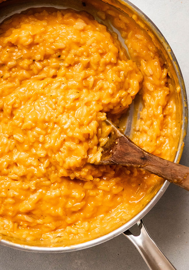Wooden spoon stirring bright orange butternut squash risotto in a wide, shallow saucepan.