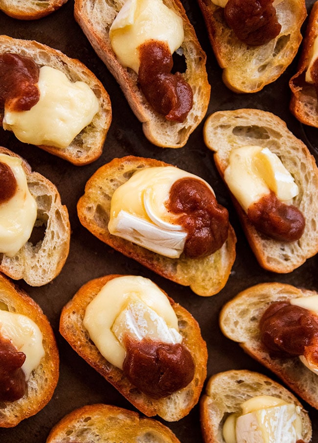 Several crostini close together on a dark background, each topped with melted brie cheese and a dollop of apple butter.