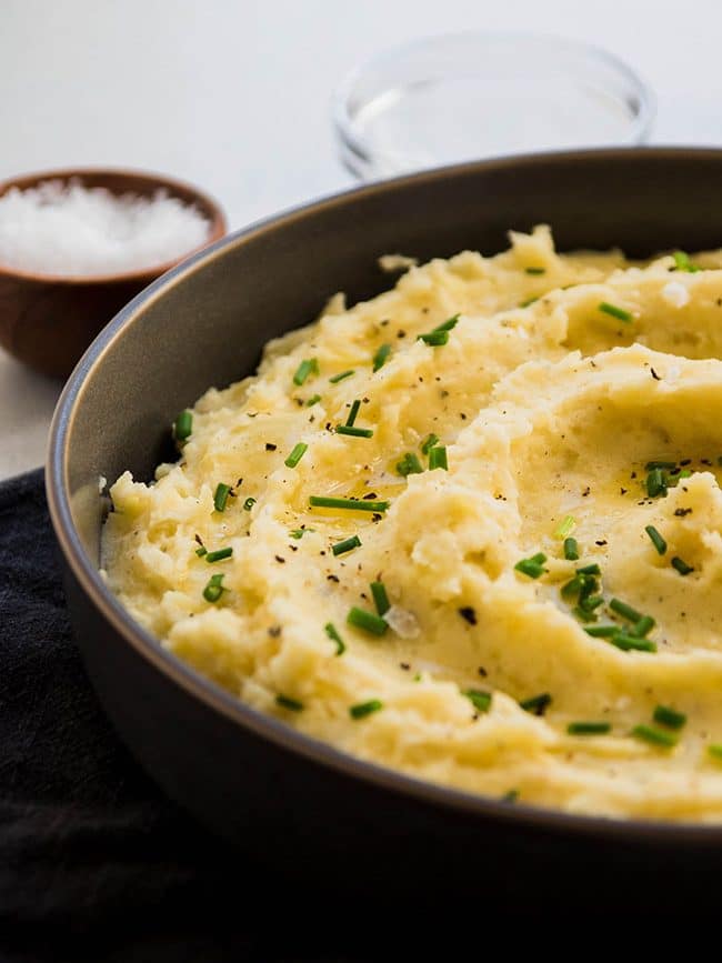 Grey bowl filled with mashed potatoes topped with fresh chives, next to a small bowl of salt.