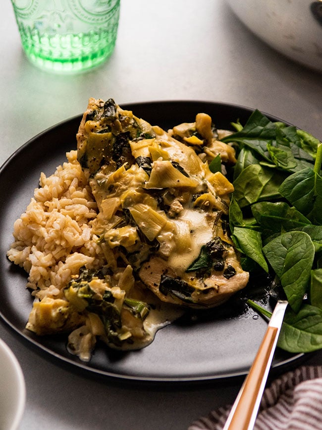 Chicken breast covered in spinach artichoke sauce on a black plate.