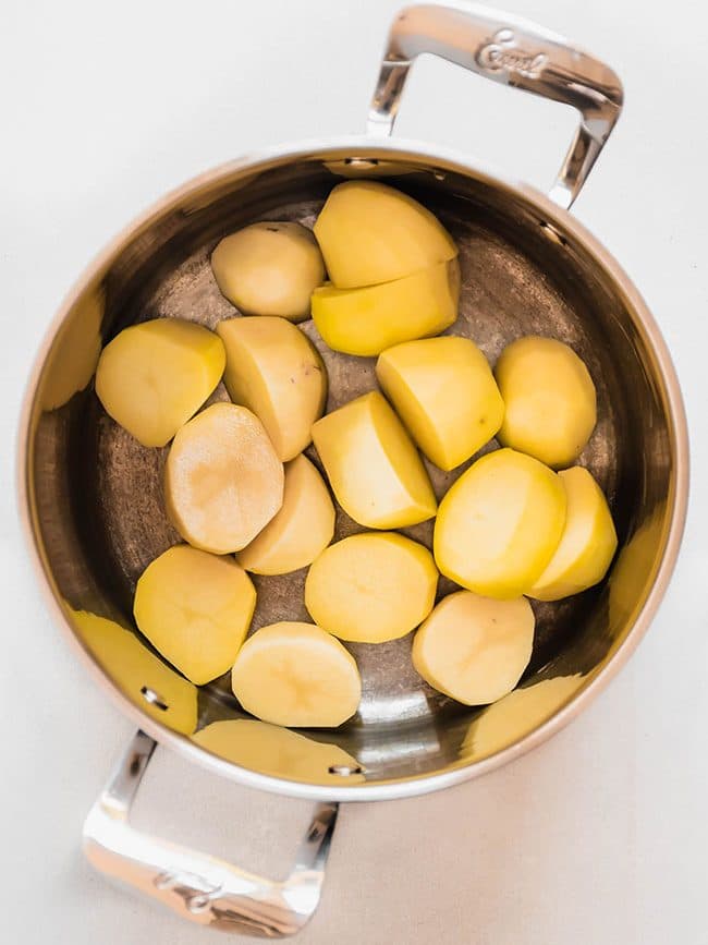 Large saucepan filled with peeled and chopped potatoes, sitting on a white surface.