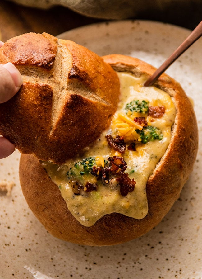 Hand dipping a piece of bread into a bread bowl filled with broccoli cheddar soup.