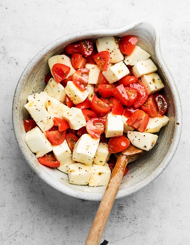 Wooden spoon stirring tomatoes and mozzarella in a large grey bowl.