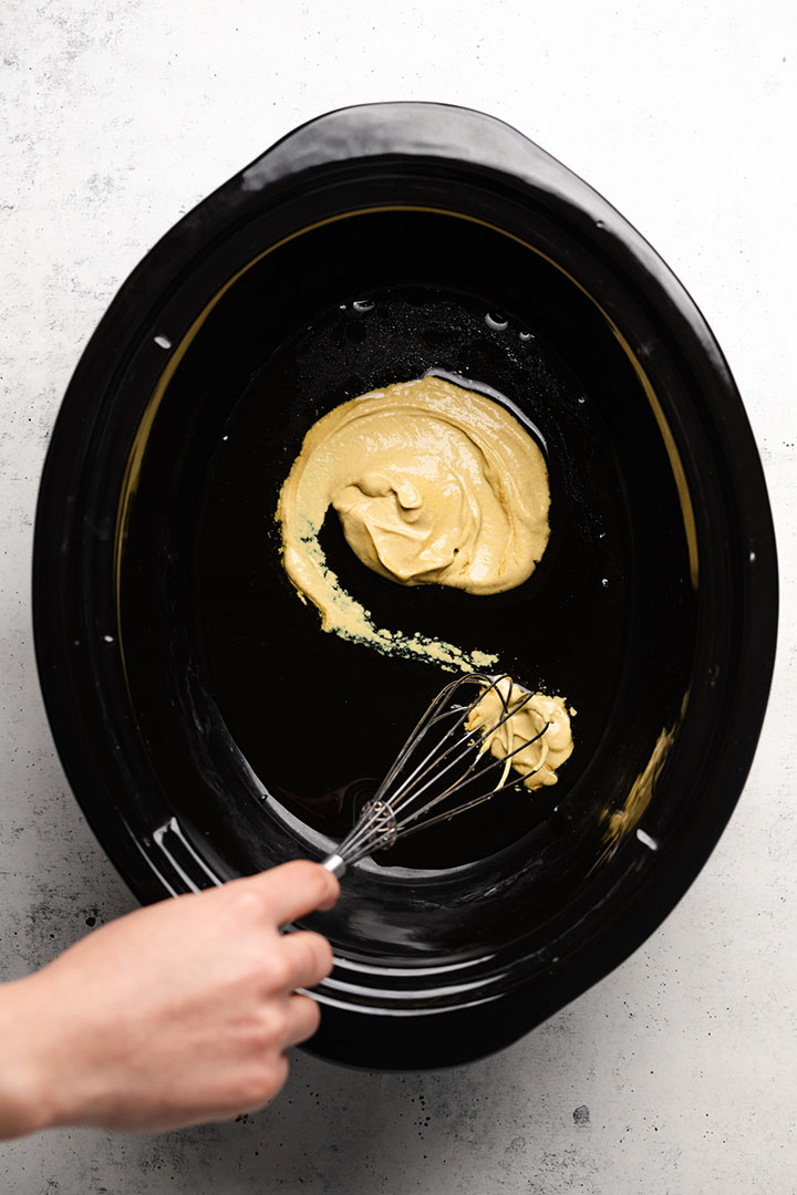 Hand whisking mustard sauce together in a black crock pot bowl.