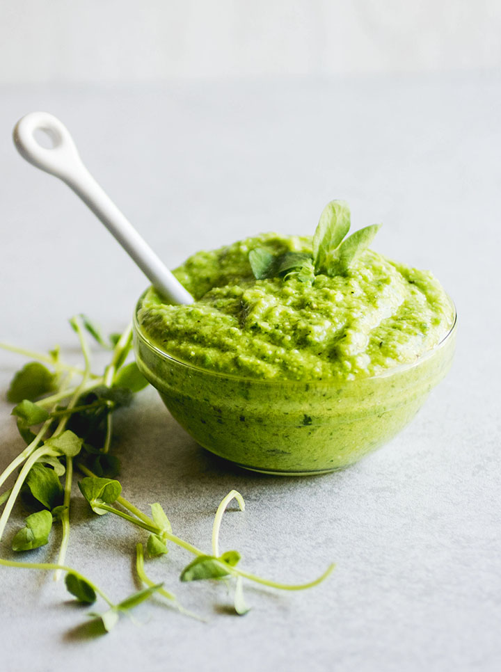 Green pesto in a small glass bowl with a white ceramic spoon sticking out of it.