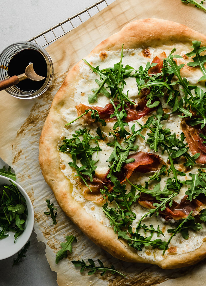 Pizza topped with fresh arugula, next to a small jar of balsamic glaze.