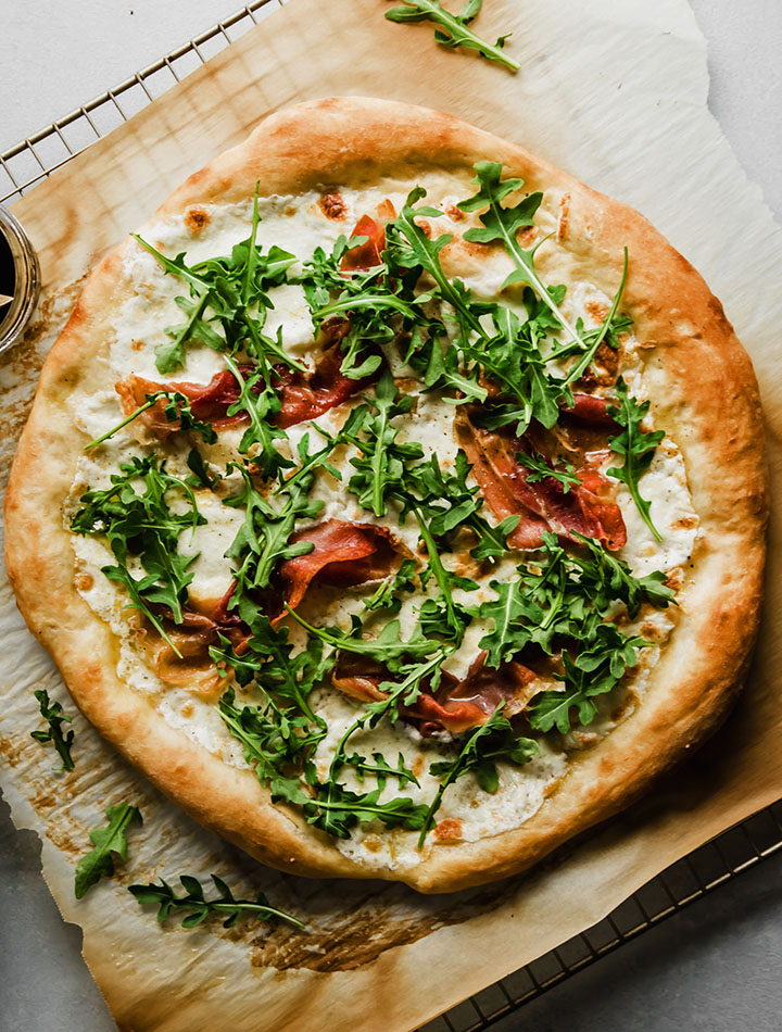 Aerial view of prosciutto pizza topped with fresh arugula, sitting on a piece of parchment paper.
