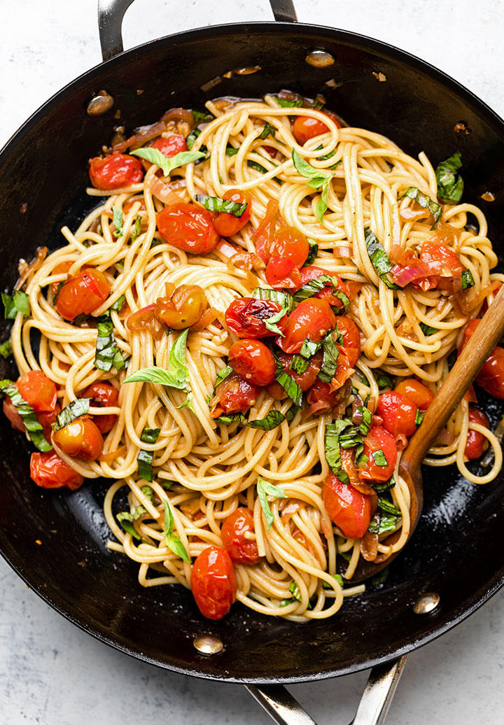 Wooden spoon stirring spaghetti and tomatoes in a black skillet.