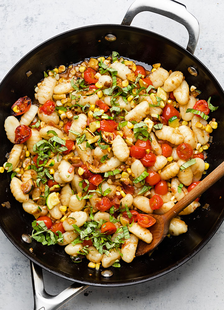 Wooden spoon stirring gnocchi with veggies in a large black skillet.