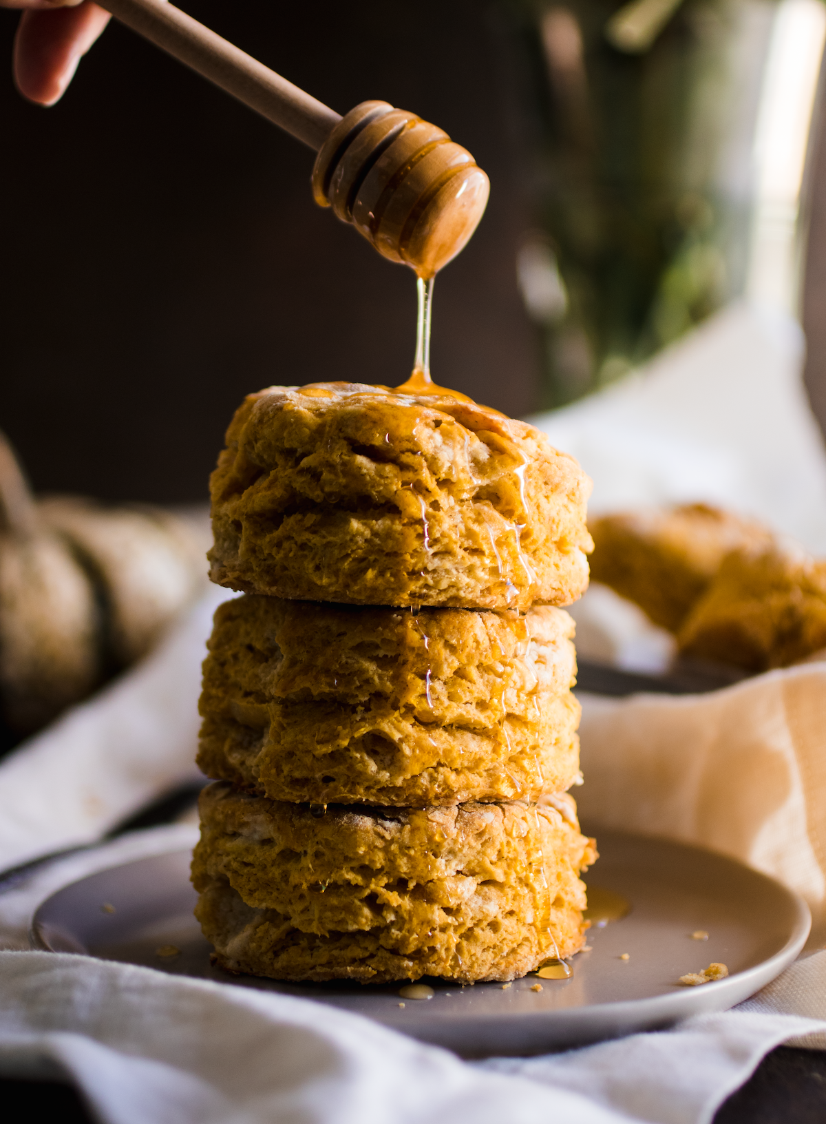 Honey dipper drizzling honey over a stack of pumpkin biscuits in front of a dark background.