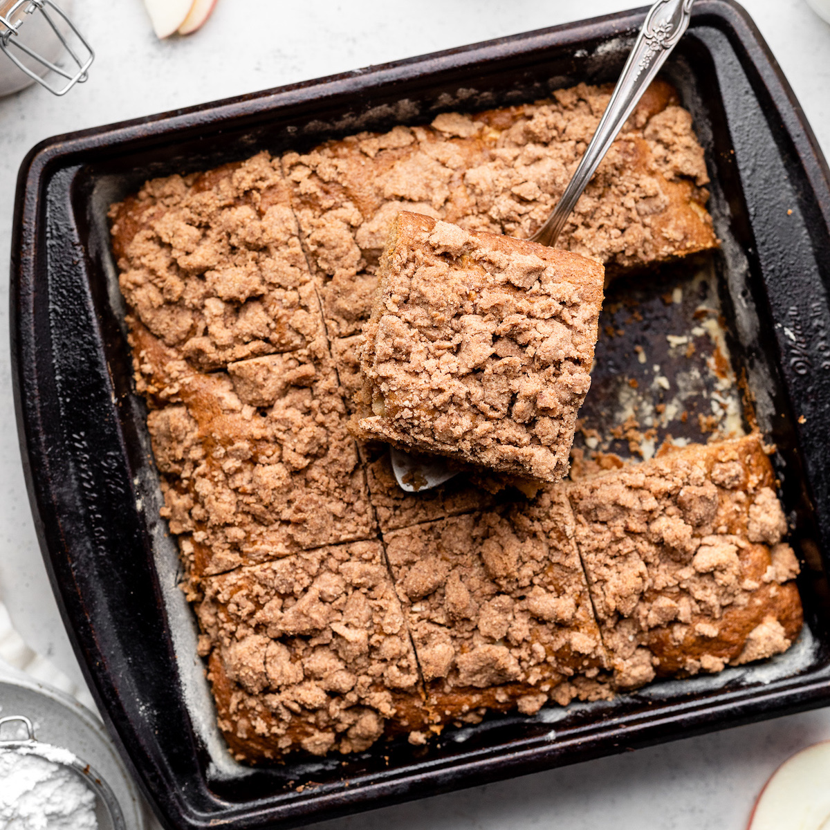 Silver serving spoon lifting a slice of coffee cake out of a square black baking pan.