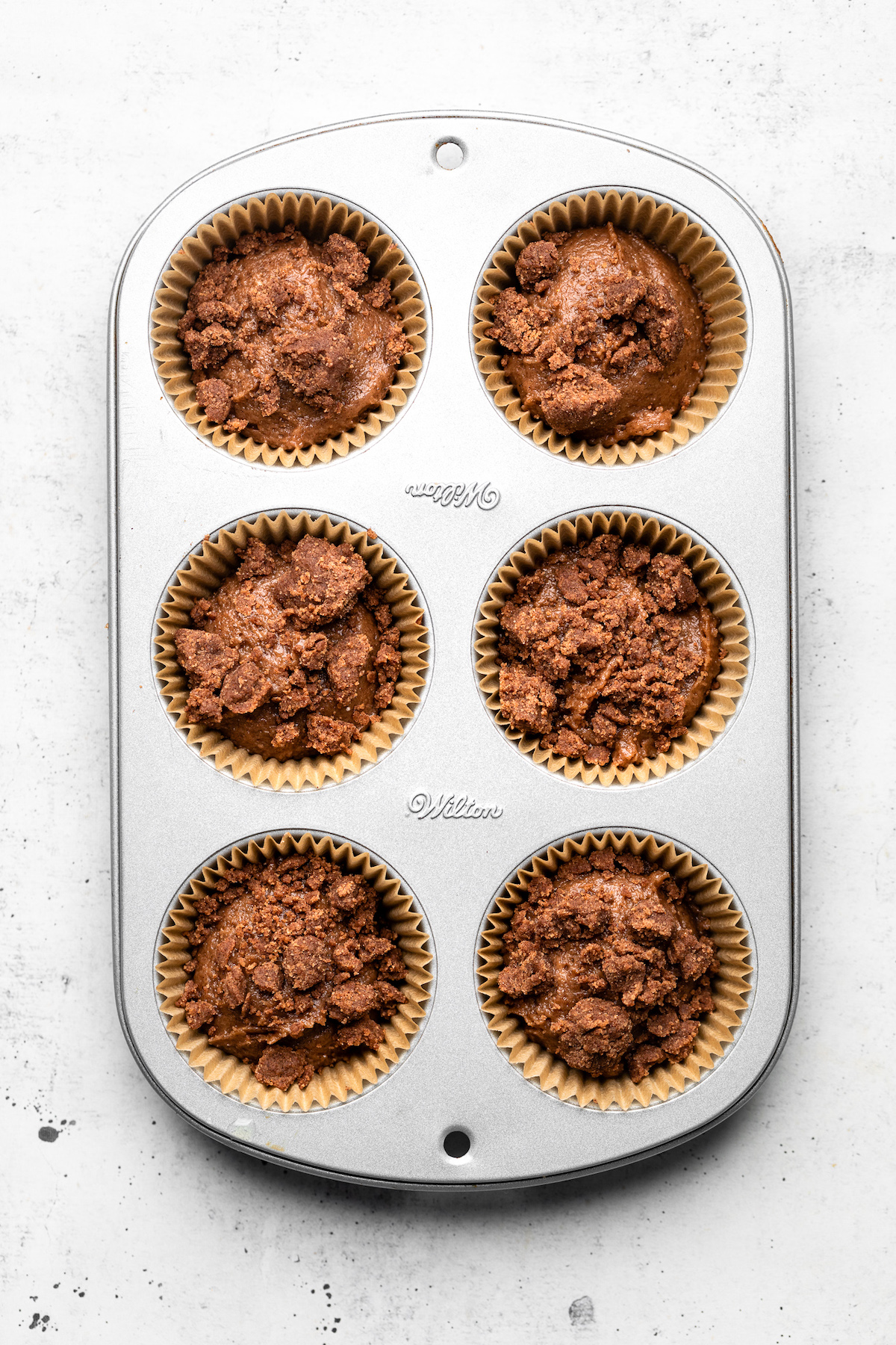 Muffin batter and crumble topping in a silver muffin tin before baking.