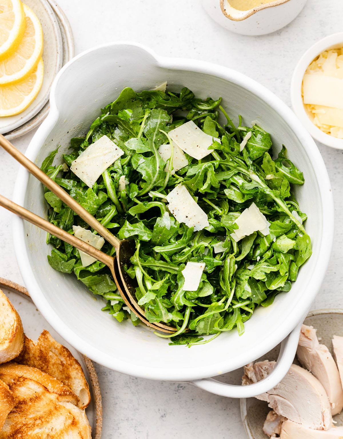 Arugula salad in a white bowl with bronze serving spoons.
