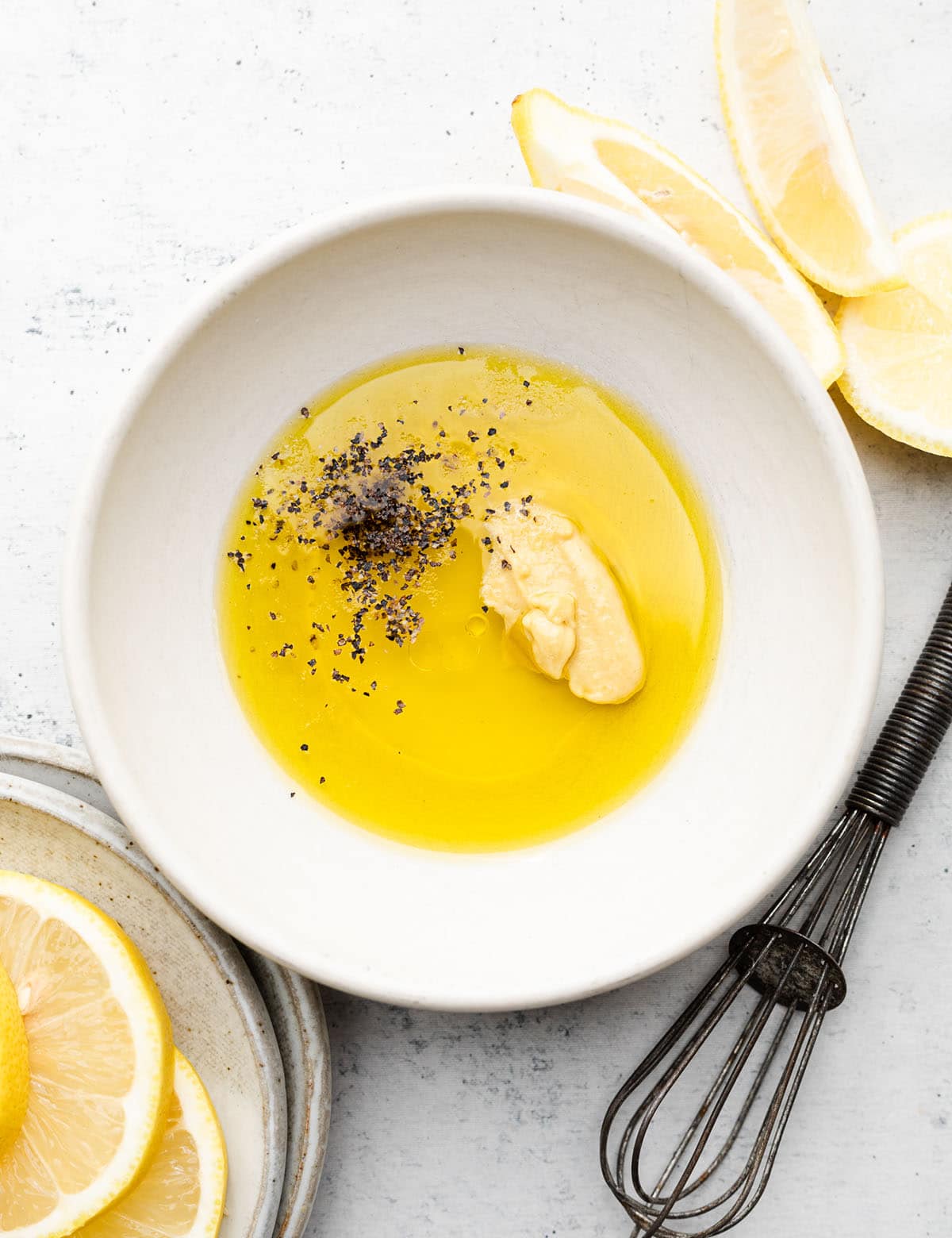 Oil, lemon juice, mustard, and pepper in a small white bowl next to a black whisk.