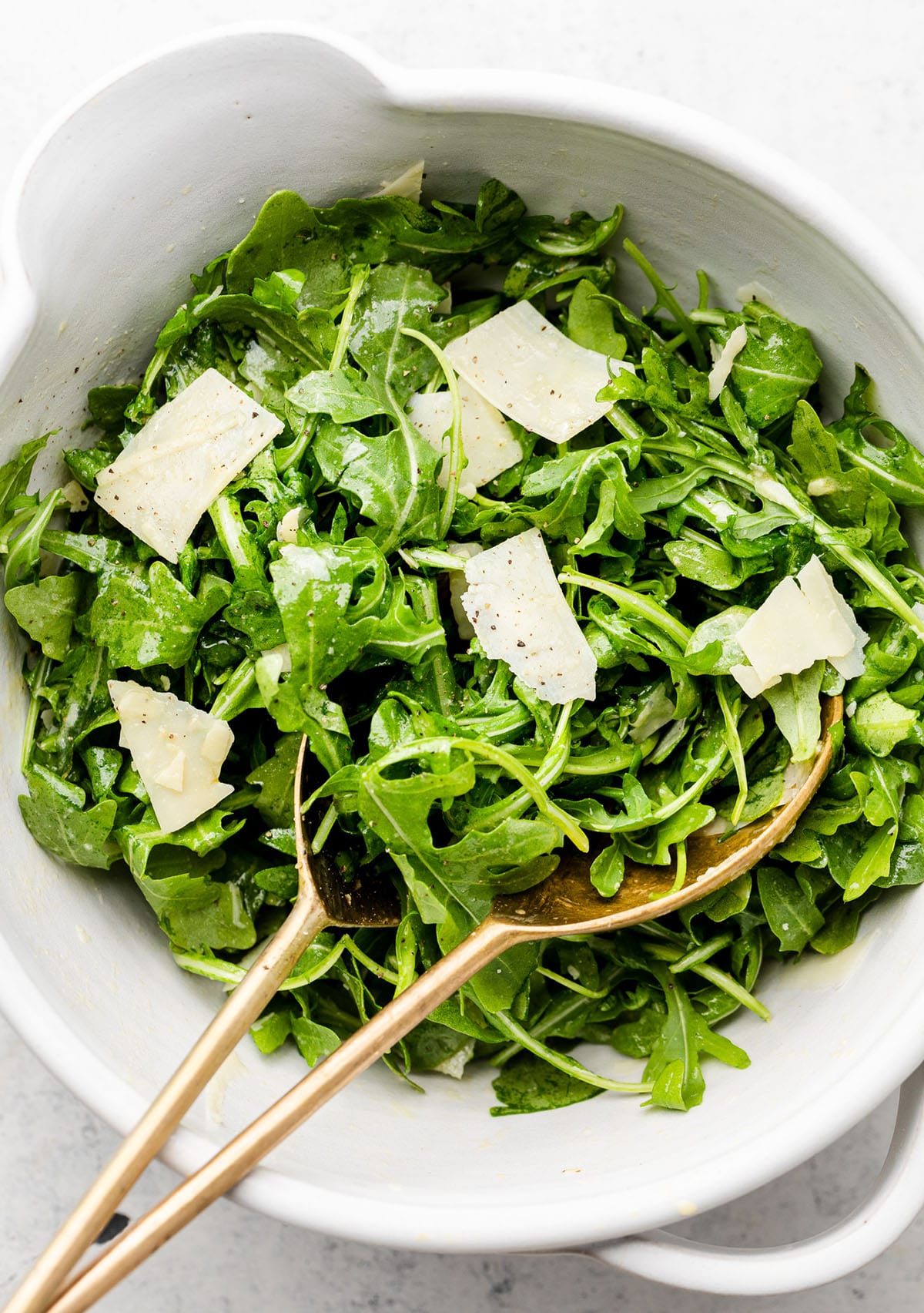 Bronze salad forks lifting a scoop of arugula salad out of a large white bowl.